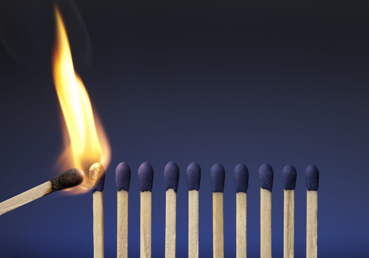 A photograph of blue-tipped matches on a blue background.  A single lit match is coming in from the left side of the image and beginning to ignite the leftmost match.  The tip of the lit match is black, and the flame is orange.  The background of the picture is deep blue at the top and gradually lightens to a brighter blue as it reaches the bottom of the image.